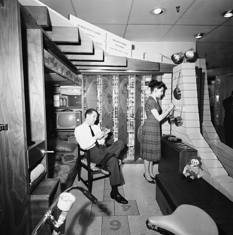 A family room designed to serve also as a fallout shelter is occupied, for demonstration purposes, by Thomas Wilson, reading, and Elise Paul, opening a can. The room is on view at the National Design Center in New York, Feb. 3, 1960 in a cutaway version to show its concrete block walls and ceiling jousts. (AP Photo/Dan Grossi)